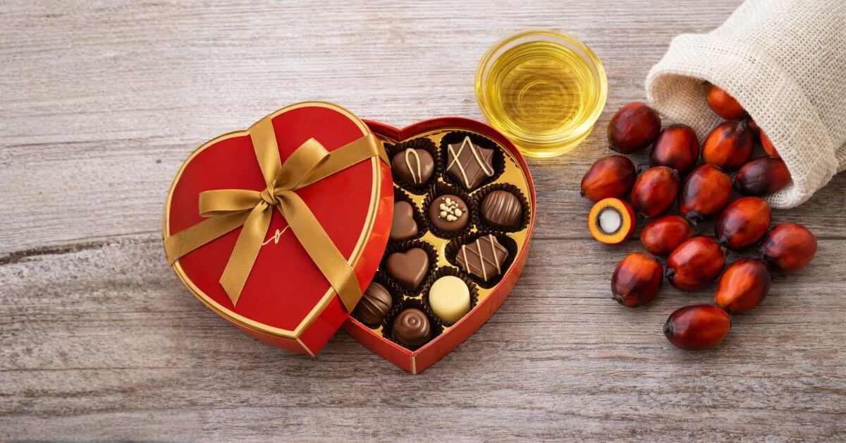 A red heart-shaped box of chocolates with a gold ribbon on a wooden table, sitting next to a burlap sack of fresh red palm fruits and a bowl of golden palm oil, representing sustainable and ethical Valentine's Day ingredients.